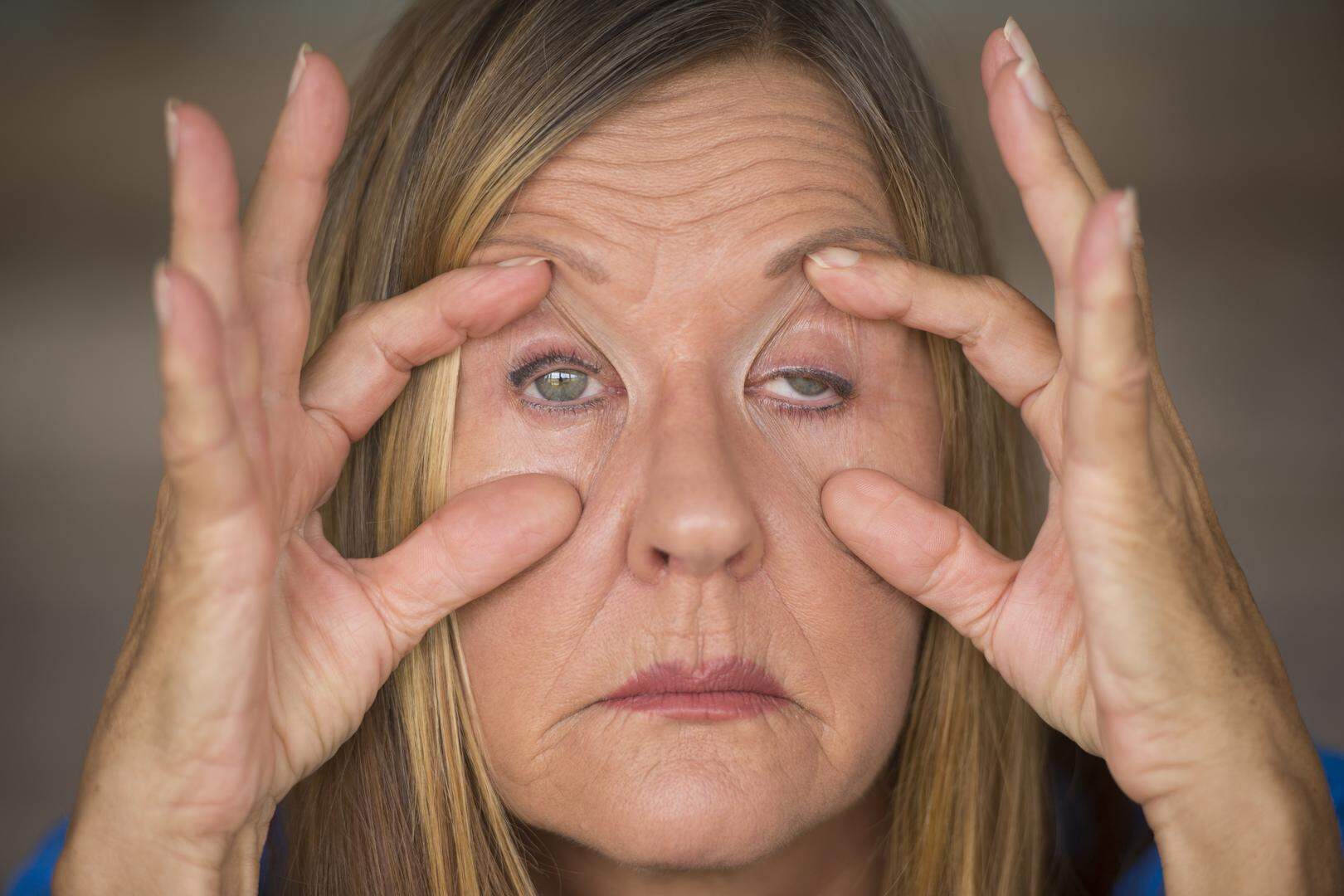 Portrait of attractive mature woman with tired and stressed facial expression, fingers keep eyes open, suffering migraine and headache, blurred background.